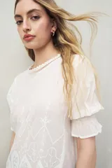 Blusa azul grisáceo de algodón con bordados frontales y terminaciones festoneadas. Tiene mangas cortas con frunces y cuello redondo con detalle de puntilla.