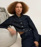 Camisa cropped de jeans azul oscuro, con cuello inglés, mangas largas, bolsillo en el pecho, pespuntes contrastantes y cierre con botones.