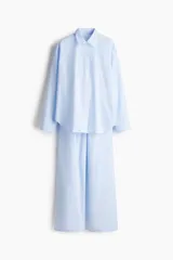 Conjunto de pijama de dos piezas en popelina de algodón a rayas celestes y blancas. La camisa es de manga larga con cuello clásico, hombros caídos y botones en delantero y puños. El pantalón tiene elástico en la cintura, cordón para ajustar y piernas amplias con vuelo en los bajos.