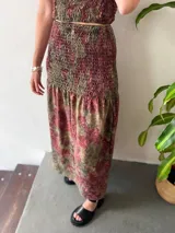 Conjunto de dos piezas compuesto por un top y una falda larga. Ambas prendas tienen un frunce elástico y un estampado floral en tonos verde oliva, rojo oscuro y negro. El top es de breteles finos y tiene espalda abierta regulable. La falda es larga y con cintura elastizada hasta la cadera.