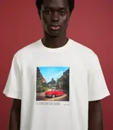 Remera de algodón color crema con cuello redondo y mangas cortas. Presenta un estampado rectangular con la imagen de un auto rojo en un paisaje y texto en la parte inferior.
