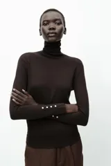 Jersey negro de punto liso con cuello alto, manga larga y detalle de botones dorados en los puños.