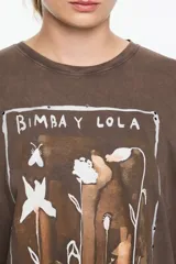 Camiseta de algodón orgánico color beige, corte oversize y manga corta ancha. Presenta un estampado central enmarcado con motivos florales y el texto "BIMBA Y LOLA" en la parte superior e inferior del diseño.