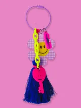 Llavero con aro metálico violeta y dijes colgantes: flor a cuadros lila y blanco con carita sonriente amarilla, cadena de eslabones verde flúor, etiqueta rosa con texto y llave rosa con carita sonriente. Incluye borla de hilos azul eléctrico.