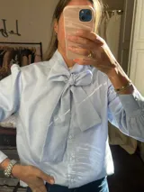 Camisa celeste con lazo en el cuello, mangas largas con puños abullonados y cierre frontal con botones.
