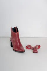 Bota corta de cuero color bordó, con taco cuadrado y punta fina. Presenta una correa con tachas pequeñas y una argolla metálica decorativa alrededor del tobillo. Cuenta con cierre lateral.