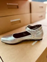 Zapato tipo Mary Jane plateado de cuero, con correa al tobillo con hebilla y tachas plateadas.