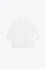 Camisa blanca de popelina de algodón, corte cuadrado, con cuello solapa, mangas cortas amplias y cierre frontal con botones ocultos.