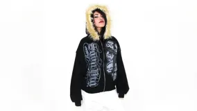 Hoodie negro con cierre, capucha con borde de piel sintética, estampado con texto y cruz con calaveras en el frente.
