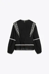 Blusa negra de algodón con cuello redondo y manga larga abullonada. Presenta bordados blancos a contraste en el cuello, hombros y mangas. Cierre en la espalda con abertura y botón.