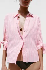 Camisa de lino a rayas verticales rosas y blancas, con cuello solapa, manga corta abullonada y lazos en los puños. Cierre frontal con botones ocultos.