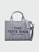 Bolso tote pequeño de cuero texturizado color gris con doble asa corta superior y correa larga extraíble y ajustable para llevar al hombro. Estampado frontal con el texto "The Tote Bag" y el nombre de la marca.