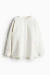 Blusa blanca de corte holgado en sarga de algodón con costuras visibles adelante y atrás. Modelo con cuello redondo, hombros caídos, tapeta con botones ocultos y mangas largas con puños abotonados. Bajo levemente redondeado con abertura pronunciada en los laterales. Espalda algo más larga.
