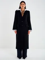 Abrigo largo negro de corte recto, con cierre frontal de un solo botón. Presenta cuello y puños blancos contrastantes, simulando una camisa debajo. Tiene bolsillos delanteros con solapa y abertura posterior.