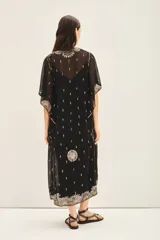 Vestido largo negro de gasa traslúcida bordada con canutillos, lentejuelas y mostacillas. Tiene escote en V, mangas amplias de largo irregular, gancho oculto en el escote y viso al tono con breteles regulables y puntilla en el escote.