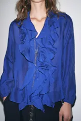 Camisa azul con escote en pico, cierre frontal con botones, mangas largas abullonadas con elástico en los puños y detalle de volantes en el frente.