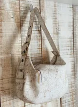 Bolso de hombro color blanco con textura craquelada, correa regulable con hebilla, interior forrado y cierre superior. Incluye llavero y bolsillos internos.