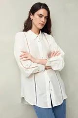 Camisa blanca de lino con botones de nácar, corte recto, cuello clásico y mangas largas con puños abotonados. Presenta detalles de calados tipo vainillas en el frente y las mangas.