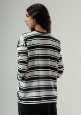 Camiseta de manga larga, corte holgado, con cuello redondo y estampado de rayas horizontales anchas en blanco y negro. Presenta un pequeño bordado rojo en el pecho.