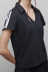 Camiseta negra tipo polo de manga corta, confeccionada en tejido de algodón. Presenta cuello clásico y escote en V, con dos rayas blancas verticales en cada manga.