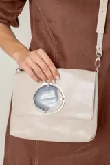 Cartera de mano tipo sobre, color beige claro, con solapa frontal decorada con una piedra de ágata azul engarzada en metal plateado. Posee correa larga ajustable de cuero beige.