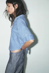 Camisa cropped de viscosa y lino a rayas verticales azules y blancas. Tiene cuello solapa, escote pico, manga corta con vuelta y bolsillos de plastrón.