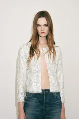 Chaqueta corta blanca con lentejuelas plateadas, cuello redondo, manga larga y forro interior.