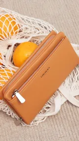 Billetera color beige con doble compartimento con cierre, correa de mano y logo de la marca en el frente.