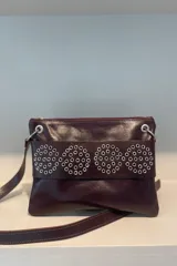 Sobre de cuero vacuno color bordó, con diseño frontal decorado con múltiples ojalillos metálicos plateados dispuestos en cuatro círculos. Cuenta con cierre superior y correa larga ajustable para llevar al hombro o cruzada.