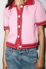 Chaqueta color blanco de punto con cuello solapa y manga corta. Tiene cierre frontal con botones color naranja y ribete en azul marino y naranja.