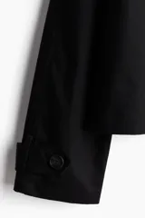 Gabardina corta negra de sarga con cuello, solapas de muesca, doble botonadura, trabilla con botón en los hombros, mangas raglán, trabilla con botón en los puños y bolsillos insertados al bies. Sin forrar.