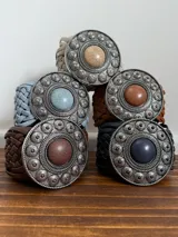 Cinto marrón trenzado con hebilla circular plateada con piedra marrón al centro.