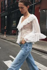Blusa blanca semitransparente de escote pico y manga larga con puño elástico. Detalle de volantes y bordados a tono. Cierre frontal con botones.
