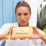 Billetera larga color amarillo pálido con textura de cuero granulado. Presenta un diseño con solapa frontal que se cierra con broche y una correa de mano desmontable. Incluye un pequeño cierre en la parte superior para el monedero y el logo de la marca grabado en la solapa.