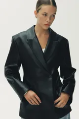 Blazer entallada negra de efecto piel, con cuello solapa y manga larga. Tiene bolsillos de plastrón en el delantero, costuras marcadas y forro interior combinado a tono. El cierre es frontal con botón interior y exterior.