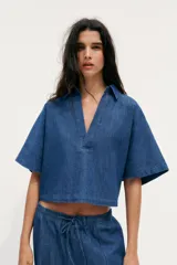 Camisa corta de denim azul, con cuello solapa, escote en pico y mangas cortas.