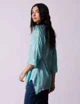 Blusa celeste con estampado de cachemir en blanco, cuello a la base, escote en V y abertura central con botones. Manga 3/4 regulable con presilla y botón, y terminación de ruedo asimétrico.