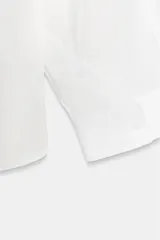Camisa blanca amplia de cuello solapa y manga larga. Tiene un bolsillo de plastrón en el pecho, bajo asimétrico y cierre frontal con botones.