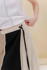 Falda midi de lino y algodón con paneles verticales en color beige, negro y tostado. Diseño portafolio con cruce, abrochada con botones y broches de presión regulables. Detalles desflecados en los cortes verticales y bolsillo escondido en el lateral derecho.