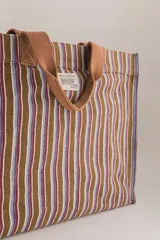 Bolso tote de algodón a rayas verticales marrones, azules y rojas, con asas de mano marrones. Tiene un bolsillo lateral interior grande, un bolsillo para el teléfono y un bolsillo para las llaves.