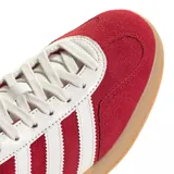 Zapatilla Adidas modelo Gazelle Indoor, con capellada de gamuza color rojo, tres franjas laterales y refuerzo de talón en cuero blanco. Posee cordones blancos y suela de goma color caramelo.