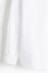 Camisa blanca de popelina de algodón con cuello inglés, tapeta clásica, dos bolsillos superiores abiertos, canesú con pliegue de caja en la espalda, mangas largas con puños abotonados, tapeta de manga con botón y bajo levemente redondeado. Corte holgado.
