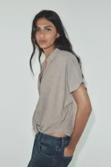Camisa color gris claro con cuello solapa y escote pico, de manga corta. Presenta un nudo en la parte delantera y cierre frontal con botones.