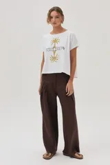 Remera blanca de algodón con cuello bote y manga corta, con un estampado central que incluye dos palmeras reflejadas y la palabra "SUNRISE" bordada en el centro.