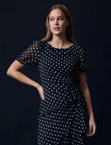 Vestido midi de gasa color negro con lunares blancos, con mangas cortas, escote redondo y ruedo asimétrico. Presenta un frunce lateral con lazo de ajuste.