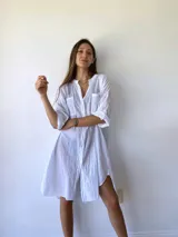 Vestido camisero blanco de algodón con cuello clásico, cierre frontal con botones, mangas tres cuartos con puños abotonados, dos bolsillos de parche en el pecho y corte holgado.