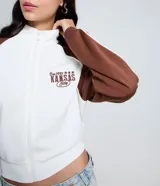Saco femenino de poliviscosa, color blanco con mangas raglán marrones y ribetes blancos. Tiene cuello alto, cierre frontal de cremallera y bordado lettering "Kansas City" en el pecho.