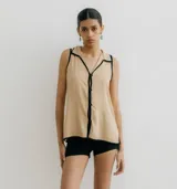 Blusa color beige con cuello camisero y botones frontales, sin mangas y con ribete negro en cuello, sisas y ruedo.