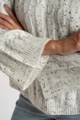 Sweater de punto color blanco roto con efecto tweed (pequeñas motas negras), cuello redondo y manga larga acampanada. Presenta un tejido texturado con trenzas verticales.