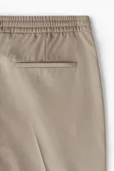 Pantalón jogger de vestir color beige, corte slim fit, con cintura elástica ajustable con cordón, pliegues delanteros y traseros, bolsillos laterales al bies y bolsillos traseros insertados.